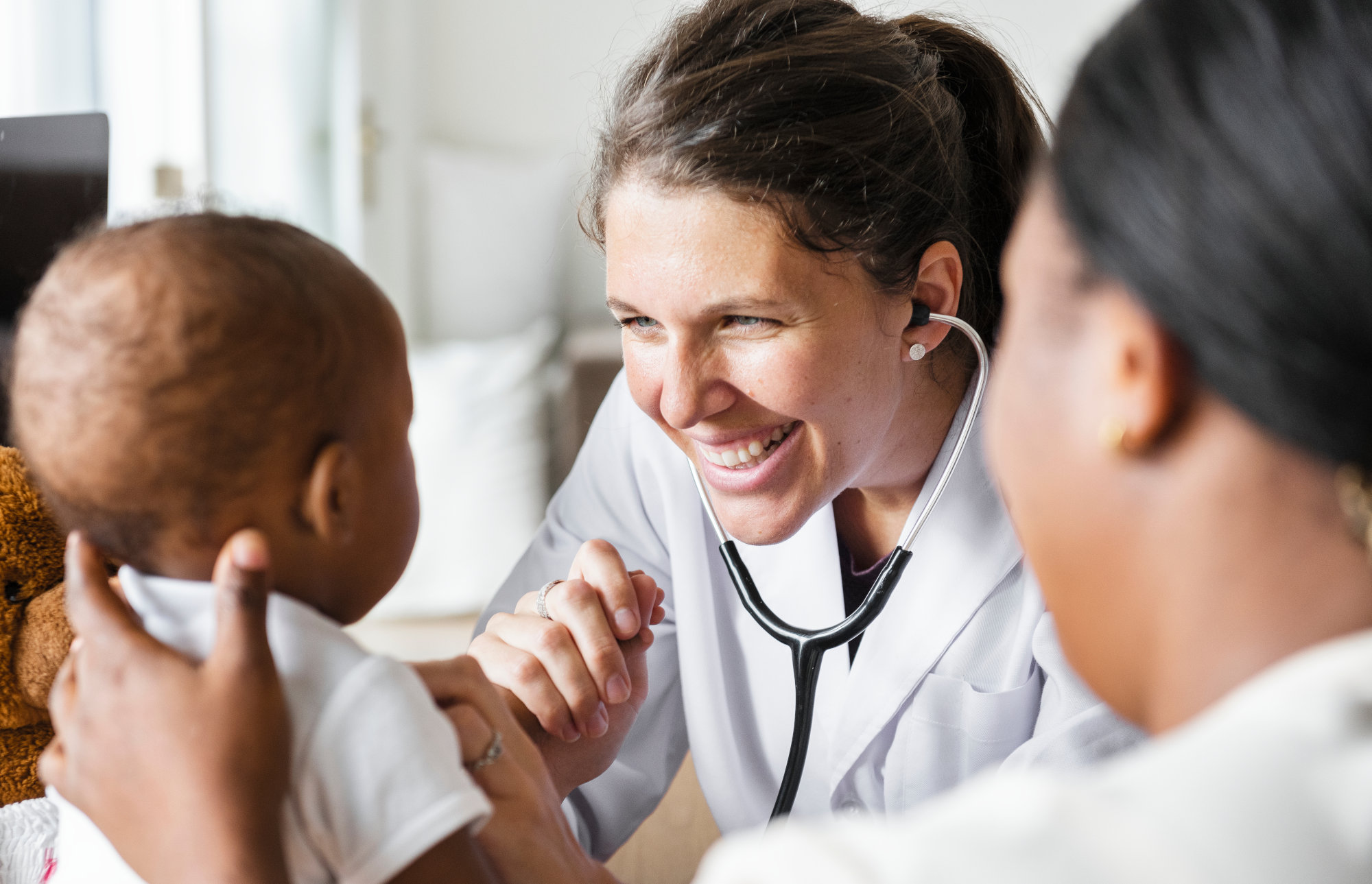 Doctor wearing a stethoscope smiling at a baby with parent alongside.