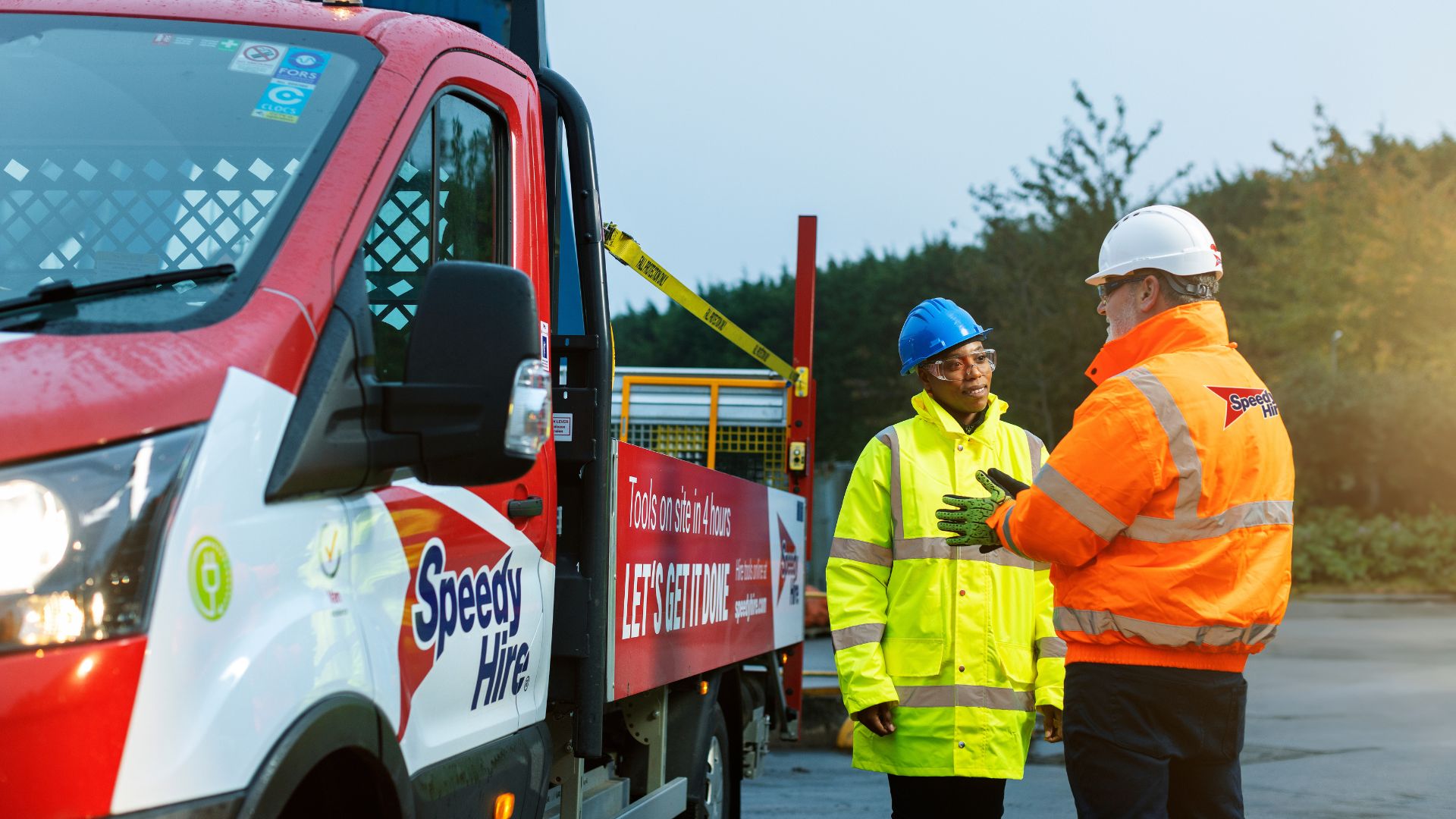 A Speedy Hire vehicle is parked beside a worksite roadway while two onsite workers wearing high‑visibility safety clothing and helmets stand next to the vehicle in discussion. The truck displays Speedy Hire branding and equipment on its rear platform, with trees and an overcast sky in the background.