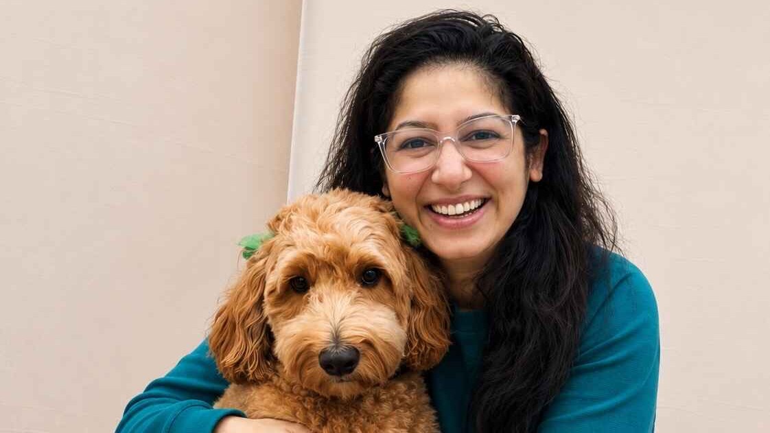 Woman wearing a teal jumper and glasses crouched next to a golden-coloured dog hugging and smiling.