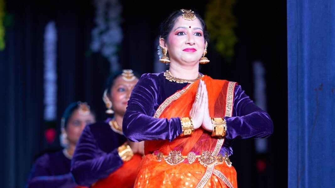 Dancer in a bright orange and purple traditional outfit performing on stage, wearing gold jewellery, with other dancers softly visible behind.