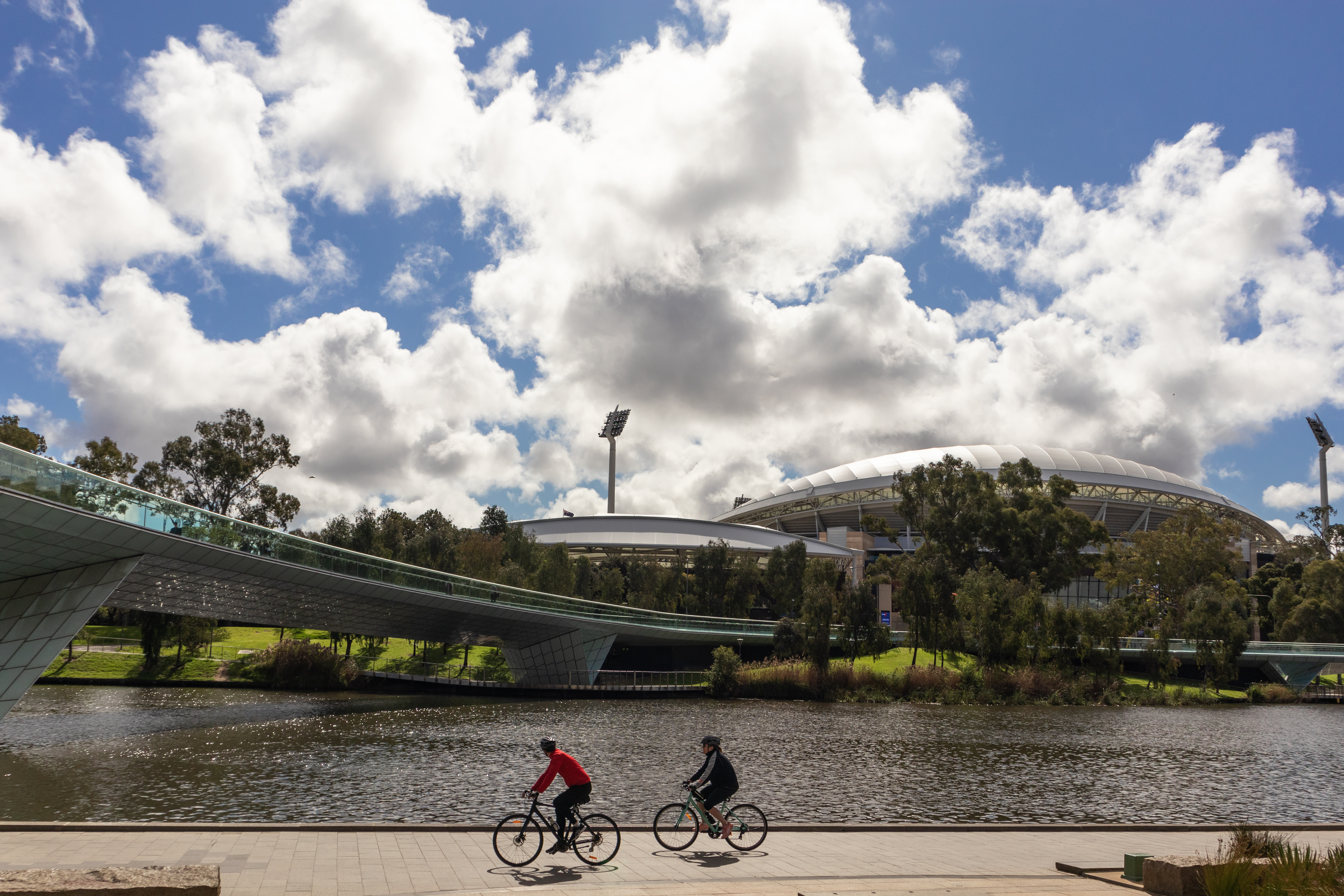 Two bikers riding near a bridge with thick white clouds in the sky. 