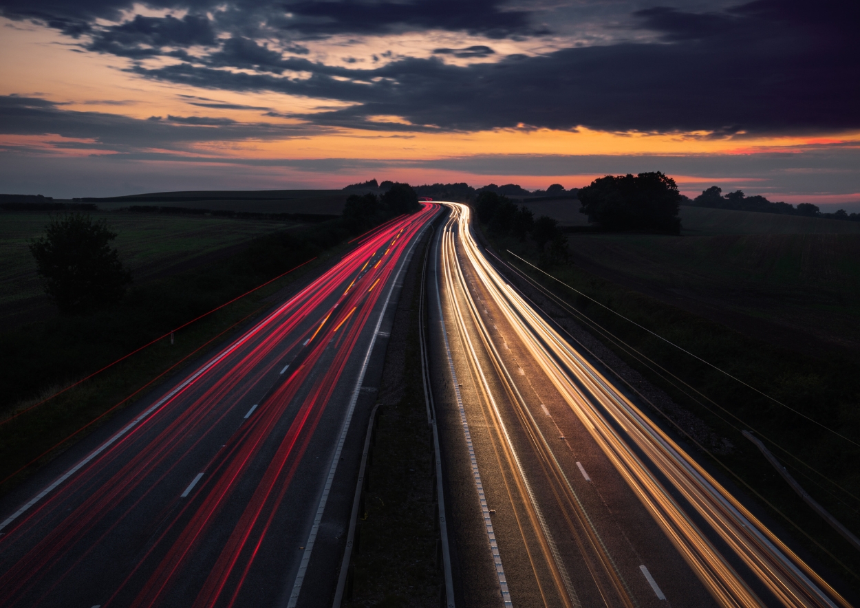 “Elevated view of the A483 motorway at dusk, with headlight trails under a vivid sunset — symbolising progress on the Welsh Government’s new infrastructure initiative.