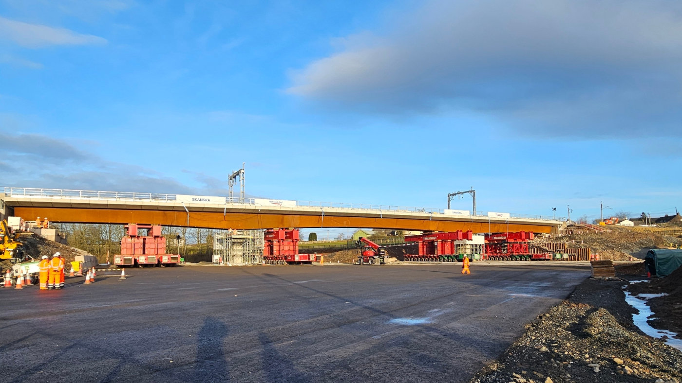 A partially constructed railway bridge in Cumbria with cranes and scaffolding in place as engineers work on the new Clifton bridge designed by Mott MacDonald. Steel beams span the river below, illustrating the progress of the structure ahead of the first trains running across it.