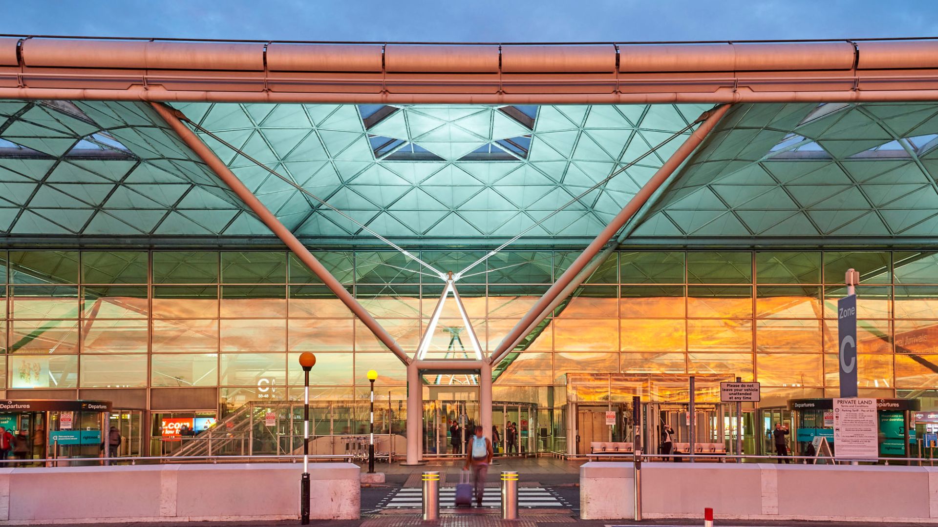 Exterior view of the Stansted Airport departures drop‑off area at sunset, showing the glass fa&ccedil;ade and distinctive roof structure with passengers walking toward the entrance.