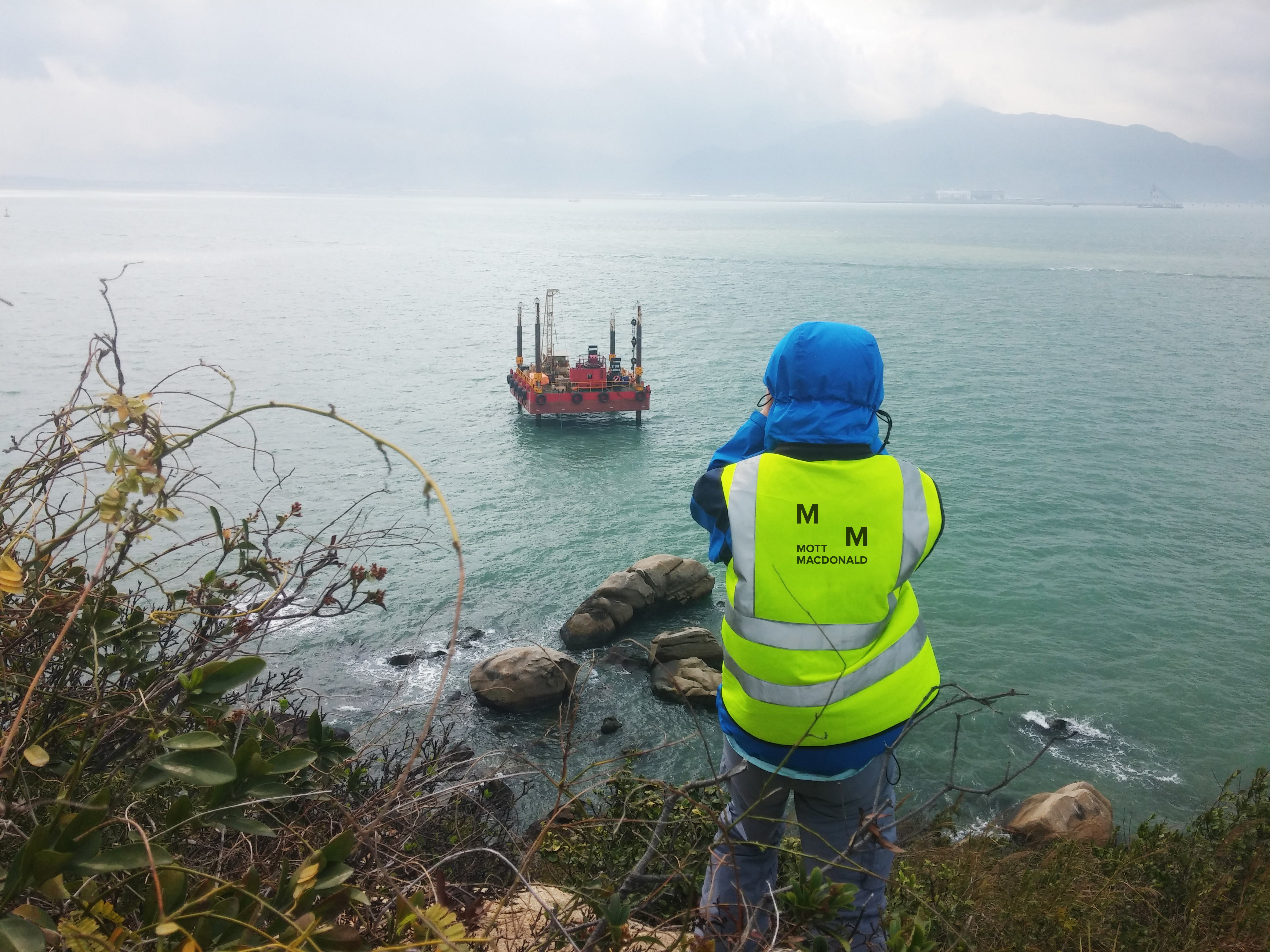 Construction worker stands in brush area looking at a vessel in the water. 
